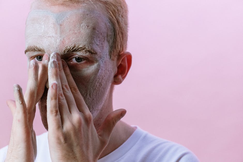Young man applying clay mask as part of his skincare routine against pink background.