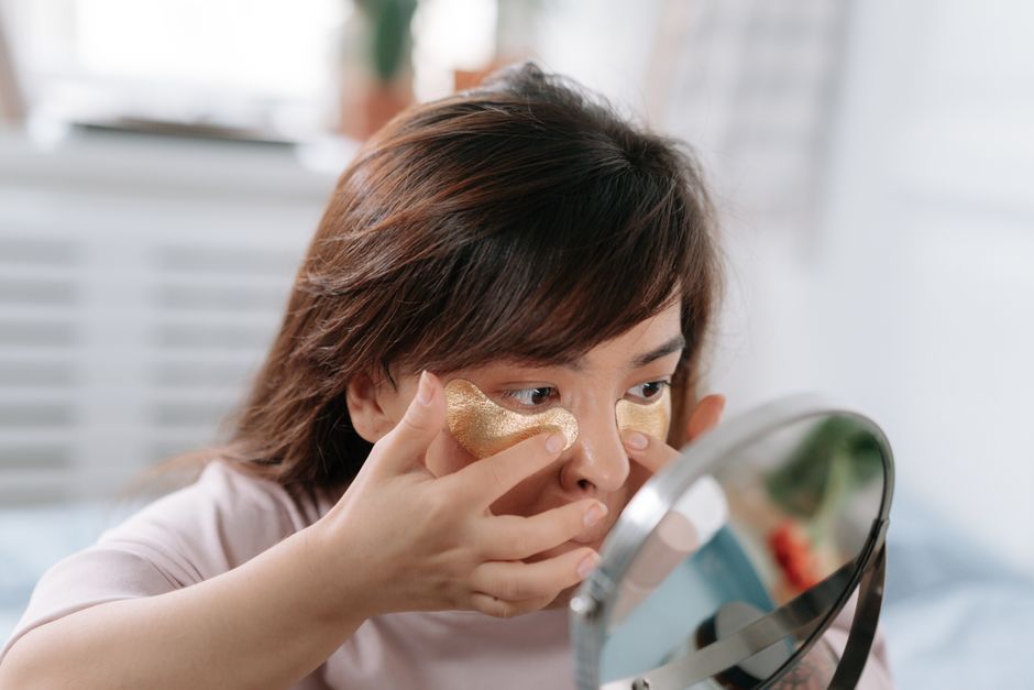 A young woman applies gold eye patches for a morning skincare routine at home.