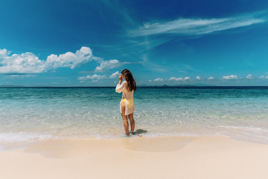 A woman stands at the edge of a tropical beach admiring the clear ocean under blue skies.