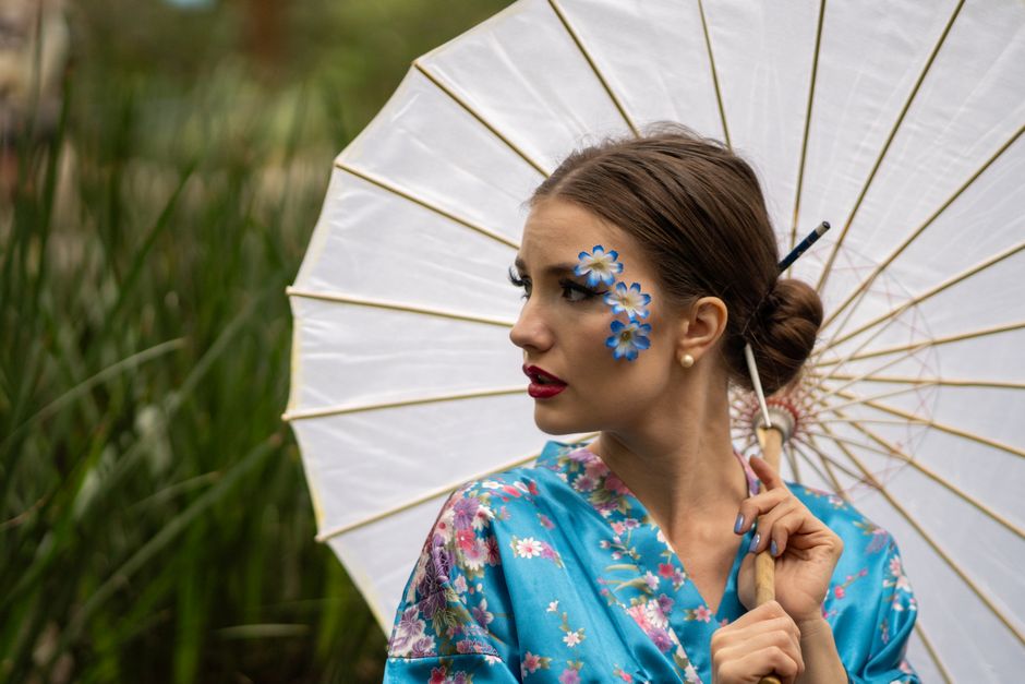A fashionable woman in a blue kimono with artistic makeup and a parasol in a park setting.