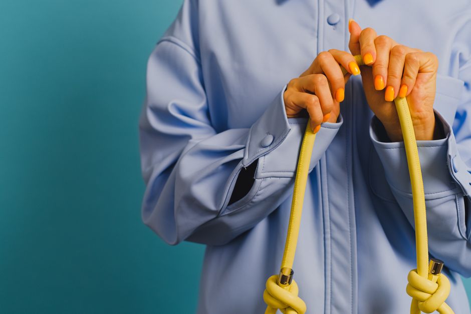 Close-up of a woman's hands with manicured nails holding a yellow rope against a light blue background.