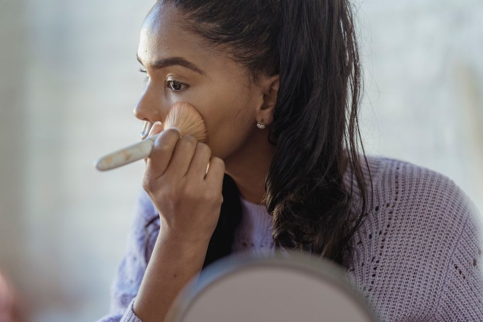 Concentrated crop African American young female spreading foundation cream on face with brush on blurred background
