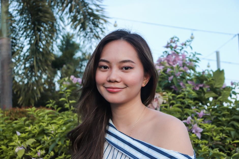 A beautiful woman with an off-shoulder top smiling amidst lush green and floral background.