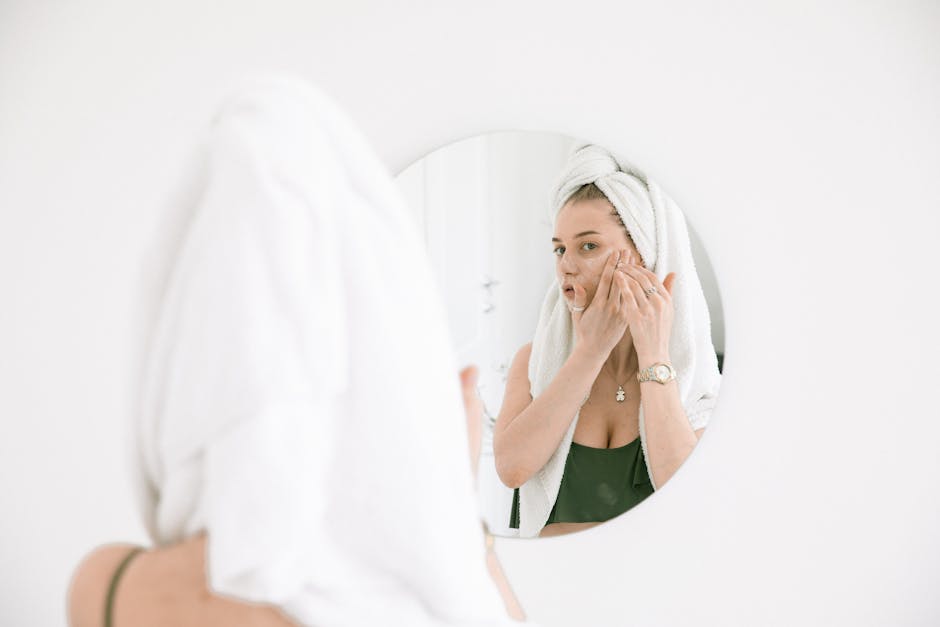 A woman applies face cream in the bathroom, reflecting in a mirror with a towel on her head.