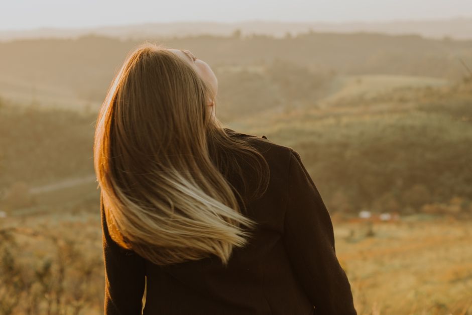 A blonde woman in a black coat enjoys the view of a serene rural landscape during sunset.