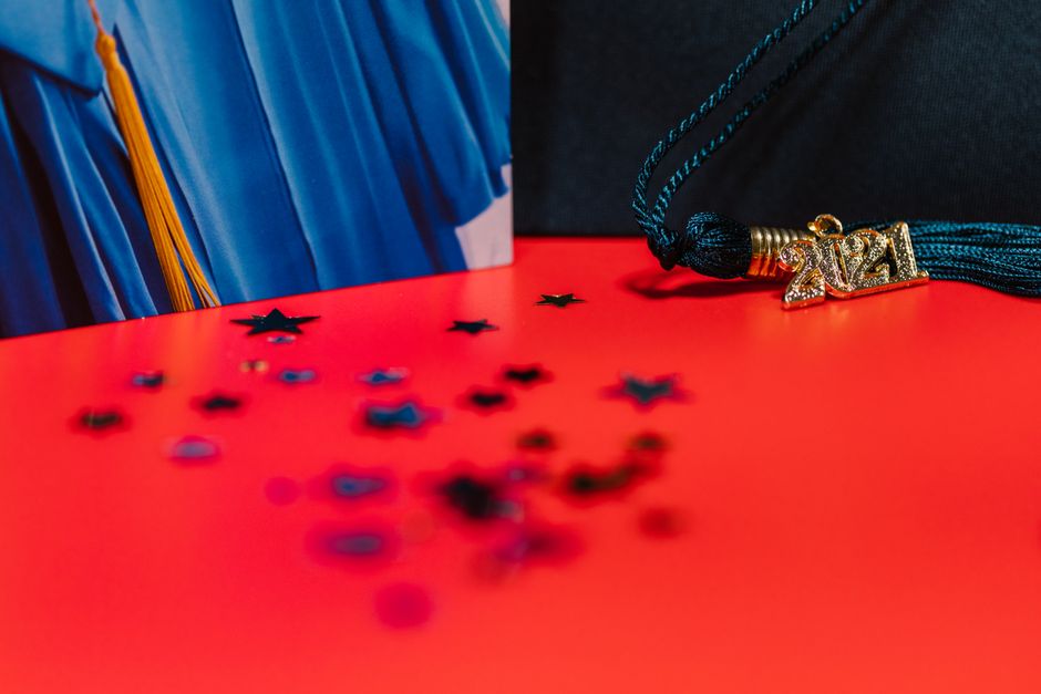 Graduation cap and tassel on bright red surface with blue gown backdrop.
