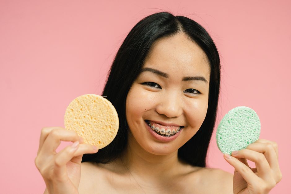 Young woman with braces holding cosmetic sponges against a pink background, smiling warmly.