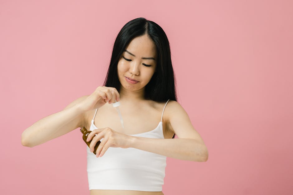 Woman using pipette to apply skincare product on her arm against pink background.