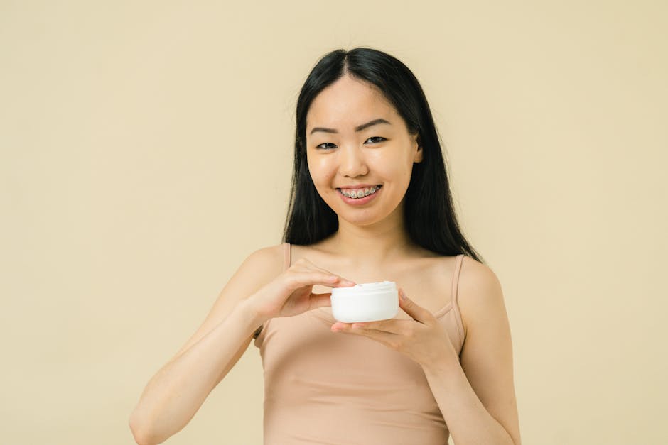 Asian woman with braces smiling while holding a cosmetic jar in a studio setting, representing skincare and self-care.