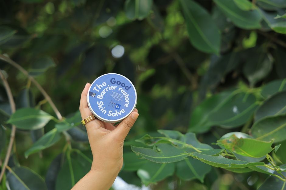A hand holds a skincare salve against a lush green leaf background.