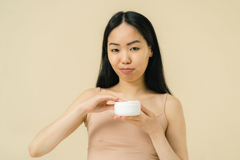 A woman with long black hair holding a cream jar in a studio setting, wearing neutral colors.