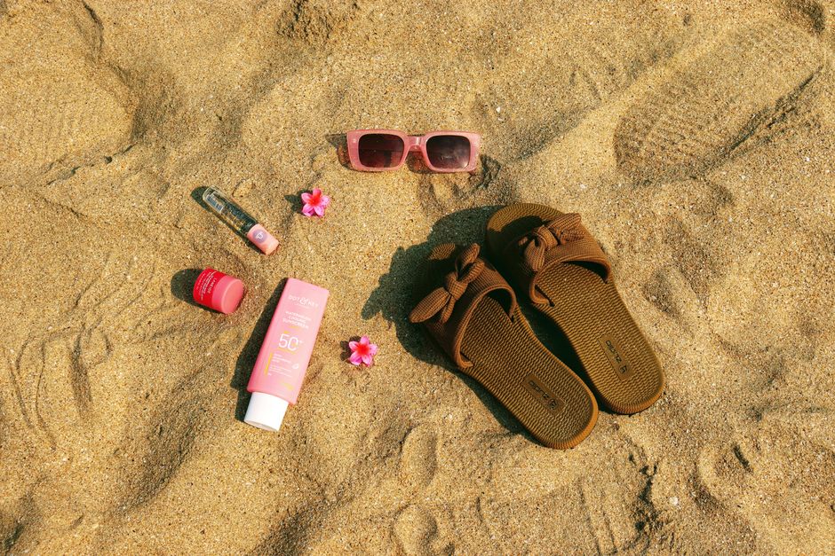 A flat lay of summer beach essentials including sandals, pink sunglasses, and sunscreen on sandy beach.